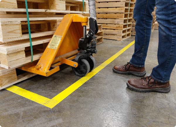 La cinta de piso ToughStripe amarilla brillante se aplica a un piso de almacén. Su posición indica donde un operador de transpaleta debe depositar un apilado de materiales.